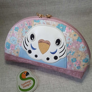 May include: A pink and blue half-circle makeup bag with a floral pattern and an embroidered parakeet face. The bag has a gold zipper and a small tag that says "Sunthaga Designs".