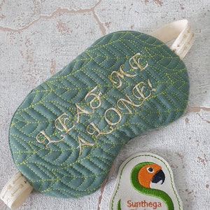 Puede incluir: Máscara de dormir acolchada verde con una correa blanca y bordado dorado que dice "I'd rather be alone!" Un pequeño parche blanco con un diseño de loro verde y naranja y el texto "Sunthaga Designs" está en la esquina inferior derecha de la imagen.