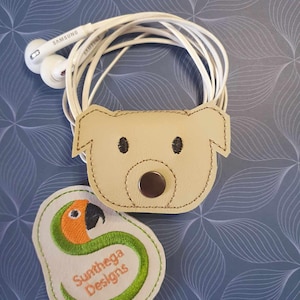 May include: A cream leather earbud tidy in the shape of a dog's face, featuring stitched black eyes and a button clasp. White earbuds are coiled around the holder. A separate patch with a parrot design and the text "Sunthega Designs" is also visible.