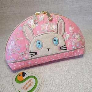 May include: A pink fabric half-moon shaped cosmetic bag with a zipper closure. The bag features an embroidered white bunny with blue eyes and black whiskers. The bag has a floral print and a pink polka dot pattern. The bag has a green and orange embroidered logo that says "Sunthaga Designs".