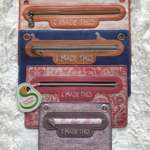 May include: Four zippered pouches with the text "I made this" embroidered on each. The pouches are made of different fabrics with various patterns and colors, including a red floral pattern, a blue fabric with a bird design, and a brown fabric with a white and orange pattern. The pouches are stacked on top of each other.