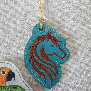 May include: A teal blue leather tag with a brown embroidered horse head design. The tag has a gold cord attached to it.