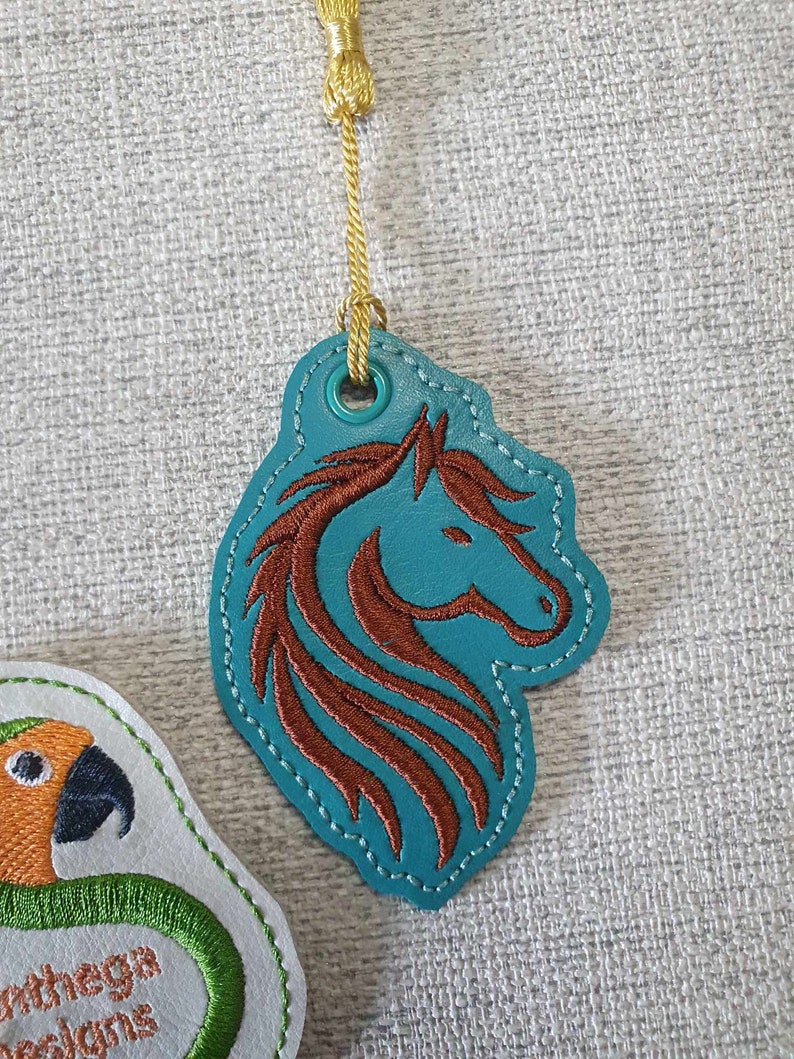 May include: A teal blue leather tag with a brown embroidered horse head design. The tag has a gold cord attached to it.