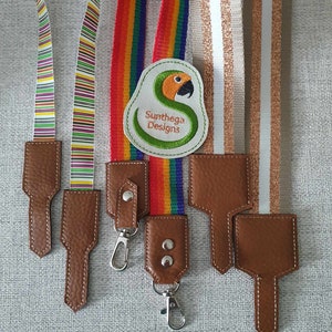 Puede incluir: Cuatro tipos diferentes de cordones coloridos con llaveros de cuero marrón. Los cordones son rayados, arcoíris, blancos con purpurina dorada y beige con purpurina dorada. Los llaveros tienen un gancho de metal y una pequeña etiqueta de cuero con el texto "Sunthaga Designs".