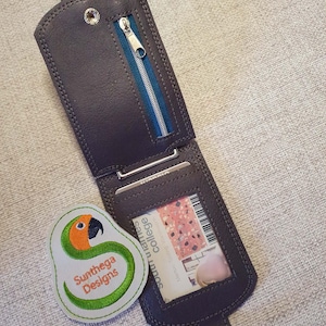 May include: Dark gray leather wallet with a zipper pocket and a clear ID window. The wallet is open, revealing a card. A small, embroidered patch with a parrot design and the text "Sunthega Designs" is next to the wallet.