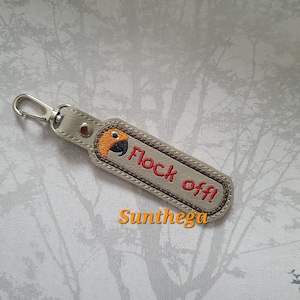 May include: A beige leather keychain with a brown stitched border. The keychain has a small embroidered bird with orange and black accents and the text "Flock off!" in brown thread. The text "Sunthega" is printed in orange below the keychain.