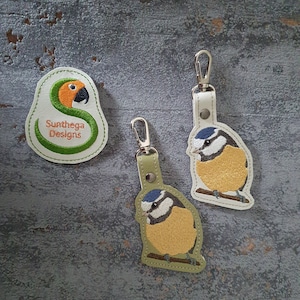May include: Three embroidered keychains and a patch featuring bird designs. The keychains depict a blue tit bird with yellow and blue plumage, perched on a branch. The patch has a parrot design with the text "Sunthega Designs".