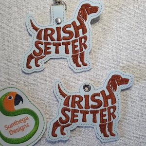 May include: Light blue Irish Setter dog-shaped keychain and tag. The dog's silhouette is filled with the words "IRISH SETTER" in brown. A small, green and white logo with a parrot is also visible.