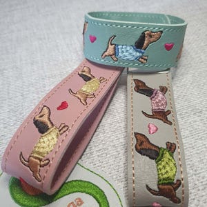 May include: Three leather key fobs in light blue, dusty pink, and light grey, each featuring embroidered dachshunds wearing sweaters and small pink hearts. A white tag with the text Sunthega Designs is visible at the bottom of the image.