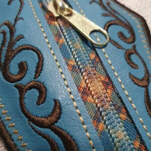 May include: A blue leather zipper pouch with a decorative brown embroidered design. The zipper is gold and has a small pull tab.