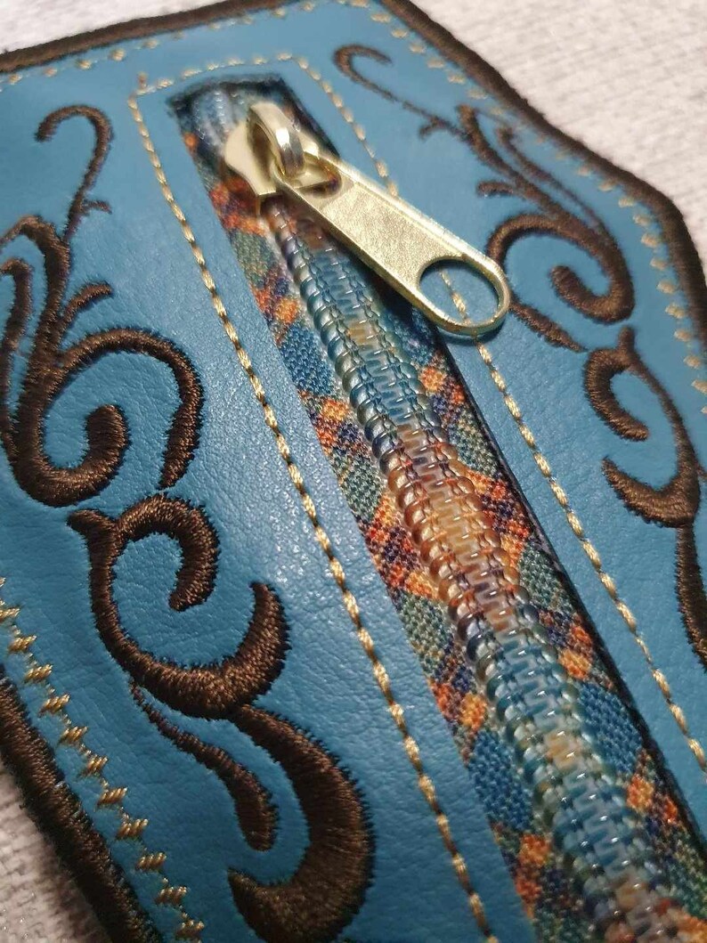 May include: A blue leather zipper pouch with a decorative brown embroidered design. The zipper is gold and has a small pull tab.