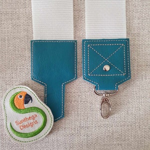 May include: White adjustable strap with teal leather accents. One end has a teal leather loop and the other end has a teal leather loop with a silver metal key ring. A white patch with a green embroidered parrot and the text "Sunhega Designs" is attached to the strap.