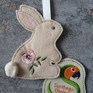 Puede incluir: Un conejito de fieltro beige con orejas rosas y una flor bordada en un lado. El conejito está sosteniendo una serpiente de fieltro verde con una cabeza naranja. La serpiente está enroscada alrededor de una etiqueta de fieltro blanco con el texto "Sunthaga Designs".