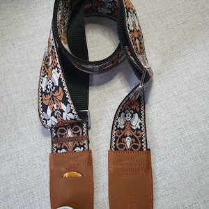 Könnte beinhalten: Ein schwarzer Gitarrengurt mit einem braunen, weißen und schwarzen Blumenmuster. Der Gurt hat zwei braune Lederenden mit einem Pickhalter an einem Ende. Der Gurt hat eine Metallschnalle.