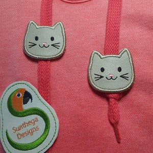 May include: A coral pink sweatshirt featuring two light gray cat face patches attached to the drawstring cords. A white patch with a green parrot illustration and the text Sunthega Designs is positioned on the lower left side of the garment.