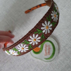 May include: A brown headband with a white daisy and green leaf embroidery. The headband is on a white surface with a green and orange logo that says "Suntega Designs".