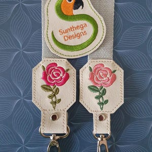 May include: Two white leather keychains with embroidered pink roses and green stems. Each keychain has a silver hook and a small white button. A beige fabric strap with a green and orange embroidered parrot and the text "Sunthaga Designs" is visible in the background.