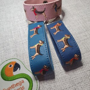 May include: A pink leather bracelet and two blue keychains, all featuring embroidered dog designs. The dogs are depicted in various colors, wearing scarves. The keychains and bracelet are accessories.