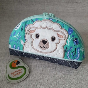 Könnte beinhalten: Eine grüne und blaue gesteppte Reißverschlusstasche mit einem weißen gestickten Lammgesicht. Die Tasche hat einen Jeansboden und ein grünes und orangefarbenes gesticktes Etikett mit der Aufschrift "Sunthaga Designs".