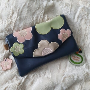 Op de afbeelding: Een blauwe leren clutch met een bloemenpatroon. De clutch heeft een klep sluiting en een klein sieraad bevestigd aan de rits. Het sieraad is een roze bloem met een wit centrum. De clutch ligt op een wit pluche oppervlak. De tekst "Sunthaga Designs" staat op een groen label.
