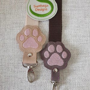 May include: Two keychains with pink paw print charms. The charms are attached to brown and beige leather straps with silver key rings. The straps have the text "Sunthaga Designs" embroidered on them.