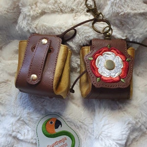 Puede incluir: Dos pequeñas bolsas de cuero marrón con detalles dorados y una rosa blanca bordada en una de ellas. Las bolsas están unidas a una cadena dorada con un cierre. Las bolsas están sobre una superficie blanca y esponjosa. El texto "Sunthaga Designs" está bordado en un logotipo de loro verde y naranja.