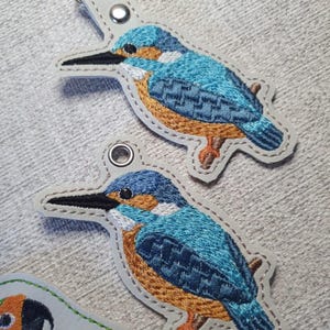 May include: Embroidered keychains featuring a blue and orange kingfisher bird design. The bird is perched on a branch, with a white border and a silver-tone keychain clasp. The bird's feathers are detailed with various shades of blue.