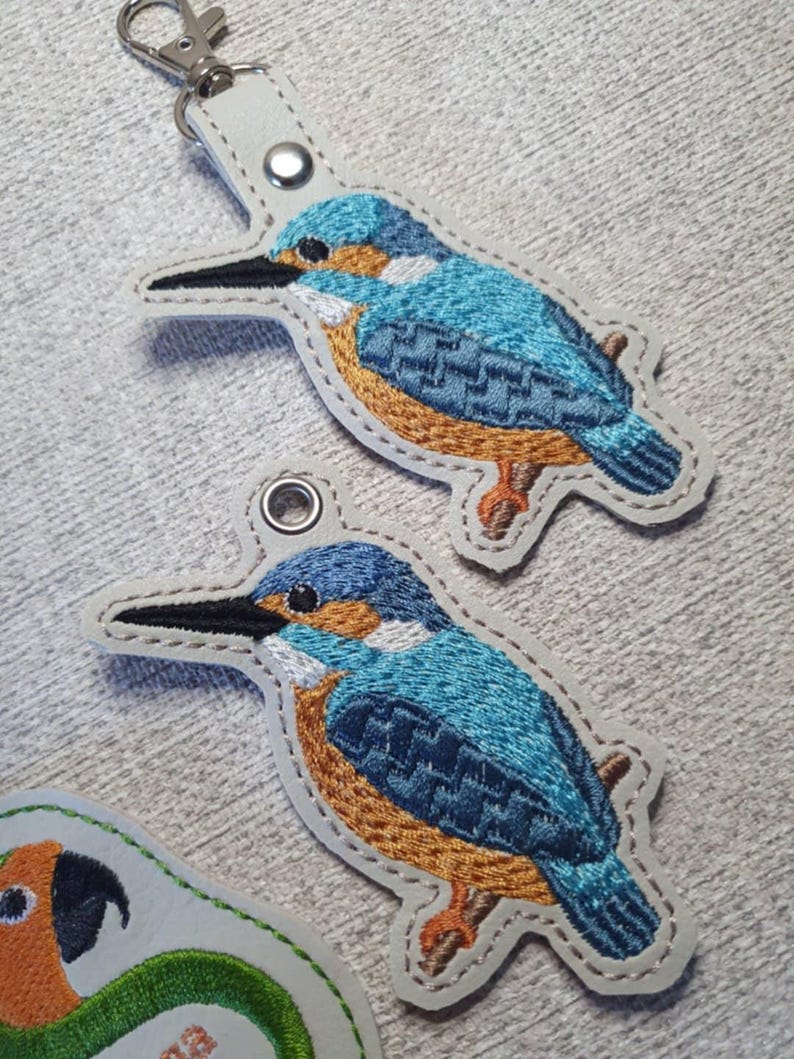 May include: Embroidered keychains featuring a blue and orange kingfisher bird design. The bird is perched on a branch, with a white border and a silver-tone keychain clasp. The bird's feathers are detailed with various shades of blue.