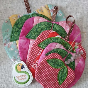 May include: Set of five quilted apple-shaped pot holders in various shades of pink, red, and green with white polka dots and gingham patterns. The pot holders have a brown ribbon loop for hanging. A small embroidered patch with a parrot and the text "Sunthaga Designs" is in the lower left corner.