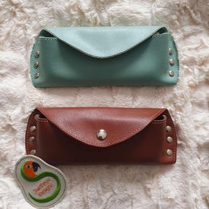 Puede incluir: Dos estuches para gafas de cuero, uno marrón y otro azul claro, con un pequeño parche bordado que dice "Sunbeaga Designs" y un diseño de loro.