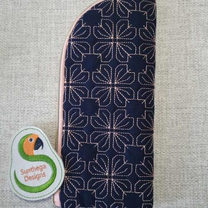 Puede incluir: Una bolsa azul marino con cremallera, forro rosa y un patrón geométrico bordado con hilo rosa. La bolsa tiene un tirador de cremallera dorado. La bolsa está sobre una superficie gris texturizada. Un parche bordado verde y naranja con el texto "Sunthaga Designs" se encuentra en la esquina inferior izquierda de la imagen.