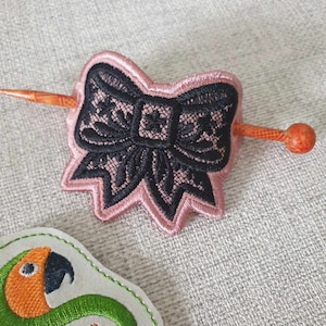May include: A black and pink embroidered bow hair clip with a wooden stick.