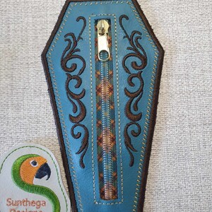 May include: A blue leather zipper pouch shaped like a coffin with brown embroidery and a gold zipper. The pouch is on a grey textured surface.
