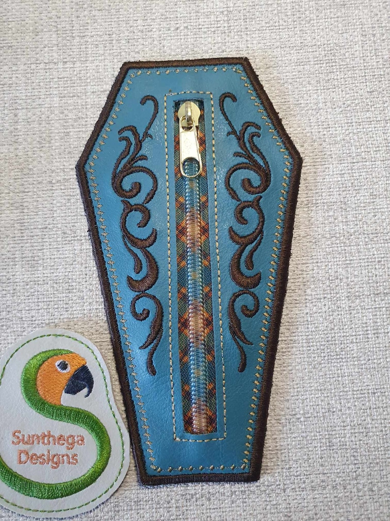 May include: A blue leather zipper pouch shaped like a coffin with brown embroidery and a gold zipper. The pouch is on a grey textured surface.