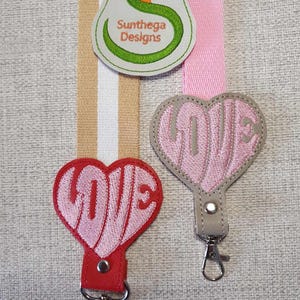 May include: Two pink heart-shaped keychains with the word "LOVE" embroidered in white. The keychains are attached to silver key rings. The keychains are on a gray background with a green and white patch that says "Sunthaga Designs".