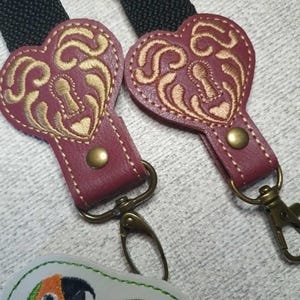 May include: Two heart-shaped, burgundy leather keychains with gold embroidered designs and brass hardware. The keychains are attached to black woven straps. A white and green embroidered parrot patch is visible.