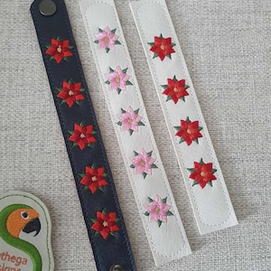 Peut inclure: Trois bracelets brodés avec un motif de poinsettia. Un bracelet est bleu marine avec des poinsettias rouges, et les deux autres sont blancs avec des poinsettias rouges et roses.
