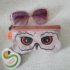 May include: Oversized, light brown sunglasses with gradient purple lenses rest above a gray owl-faced glasses case. A small, white patch with a green and orange parrot design and the text "Sunthega Designs" is also visible.