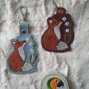 May include: Two embroidered fox keychains with a blue and a maroon background. The foxes are both looking at a white flower. The keychains are hanging on a white fuzzy surface. A small white patch with green embroidery and the text "Sunthaga Designs" is in the lower right corner of the image.