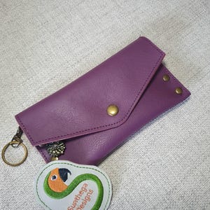 May include: A purple leather wallet with a flap closure and brass-colored rivets. A keychain and a white patch with a parrot design and the text "Sunthega Designs" are also visible.