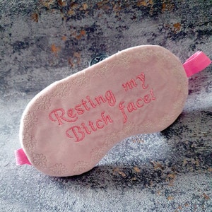 Puede incluir: Máscara de dormir rosa con bordado floral blanco y el texto "Resting my Bitch face!" bordado en rosa.
