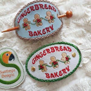 May include: Two embroidered patches with the text "Gingerbread Bakery" and three gingerbread men holding hands. The patches are on a white fluffy surface.