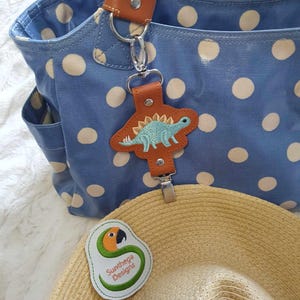May include: A blue polka dot tote bag with a brown leather handle and a dinosaur-shaped bag charm. The charm is attached to a silver clip. A straw hat with a green and orange parrot patch that says "Sunthaga Designs" is hanging from the clip.