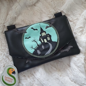 May include: Black clutch bag with a Halloween embroidery. The design shows a haunted house, bats, and a graveyard scene in a circular frame. The bag has a flap closure and a shoulder strap.