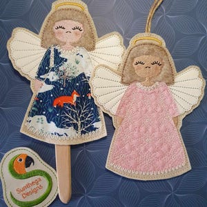 May include: Two felt angel ornaments, one with a blue and white winter scene and a fox, the other in pink. Both have gold haloes and white wings. The angel with the winter scene is attached to a wooden stick.
