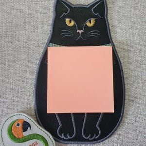 May include: A black cat-shaped memo holder with yellow eyes and a pink sticky note. The holder has a blue and green collar. A small, white, avocado-shaped patch with a parrot design and the text "Sunthega Designs" is next to the memo holder.