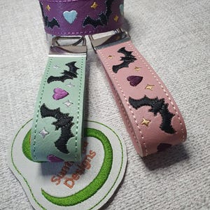 Puede incluir: Tres llaveros con murciélagos negros, corazones y estrellas bordados. Uno es morado, otro verde menta y otro rosa empolvado. Cada uno tiene un cierre de metal plateado. Un parche de cuero sintético blanco con el texto Sunn Designs bordado.