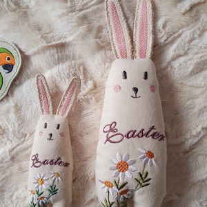 Könnte beinhalten: Zwei bestickte Plüsch-Hasenspielzeuge mit rosa Ohren. Der größere Hase hat das Wort "Ostern" auf seiner Brust gestickt und weiße Gänseblümchen mit orangefarbenen Zentren. Der kleinere Hase hat das Wort "Ostern" auf seiner Brust gestickt.