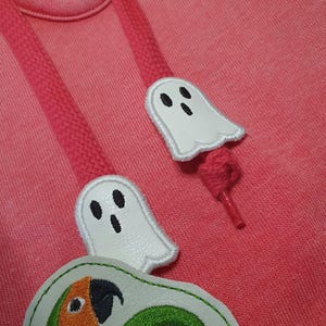 Puede incluir: Una sudadera con capucha de color rosa coral que presenta dos parches de fantasmas blancos con ojos y bocas bordados en negro, unidos a los cordones rosas. Un tercer parche muestra un diseño de loro verde y naranja con texto parcial.