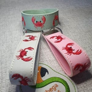 以下が含まれることがあります： 赤いカニの刺繍が施された3つのリストレットキーホルダー。ミントグリーン、白、ライトピンクの3色です。各キーホルダーにはシルバーカラーの金属リングが付いており、Sunthega Designsという文字が入った白いパッチの近くに配置されています。